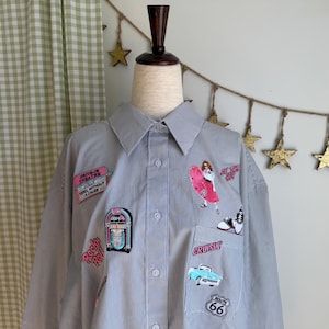 Puede incluir: Camisa a rayas azules y blancas con parches bordados. Los parches incluyen una gramola, un coche clásico, una mujer con una tabla de surf y el letrero de la Ruta 66. La camisa se exhibe en un maniquí.