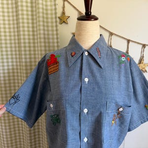 May include: A light blue button-up shirt with embroidered designs of a spider, frog, snake, and other floral and insect motifs. The shirt has two front pockets and a collared neckline.