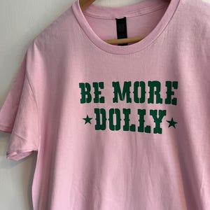 May include: A light pink short-sleeved t-shirt featuring the dark green text "BE MORE DOLLY" centred on the chest. The text is written in a bold, western-style font and is flanked by two small dark green stars on either side of the word DOLLY.