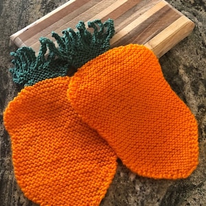 May include: Two orange knitted pot holders with green fringe on top. The pot holders are shaped like carrots.