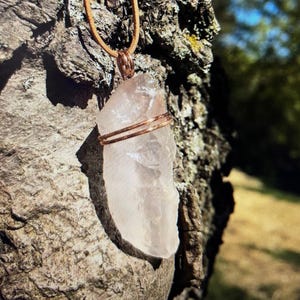 May include: A clear quartz crystal pendant wrapped in copper wire, hanging from a brown leather cord. The pendant is hanging against a tree trunk.