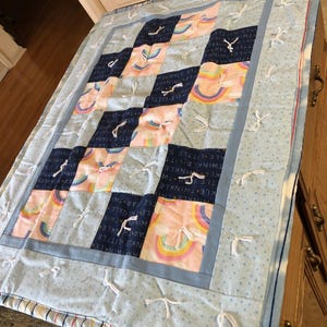 May include: A blue and pink baby quilt with a rainbow pattern and the words "Twinkle Twinkle Little Star" repeated throughout. The quilt has white ties on each square.