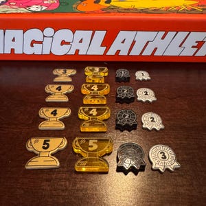 May include: A collection of game pieces, including gold and silver trophy-shaped tokens with numbers 3, 4, and 5, and black and white ribbon-shaped tokens with numbers 1, 2, and 3. The game box is visible in the background.