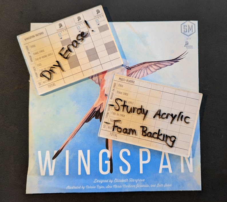 May include: A close-up of the Wingspan board game box with two dry-erase score sheets. The score sheets are made of sturdy acrylic with a foam backing. The text on the score sheets reads "Dry Erase" and "Sturdy Acrylic Foam Backing".