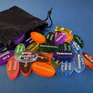 May include: A black drawstring bag spilling out a collection of colourful, oval-shaped game pieces. Each piece is transparent and features a character name in white text, such as "Evil Queen" and "Captain Hook". The pieces are scattered on a blue surface.