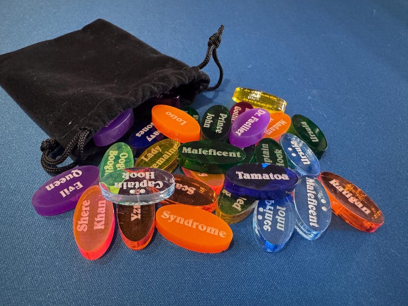 May include: A black drawstring bag spilling out a collection of colourful, oval-shaped game pieces. Each piece is transparent and features a character name in white text, such as "Evil Queen" and "Captain Hook". The pieces are scattered on a blue surface.