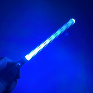 Peut inclure: Une baguette lumineuse bleue avec une lueur blanche. La baguette est tenue dans une main sur un fond bleu.