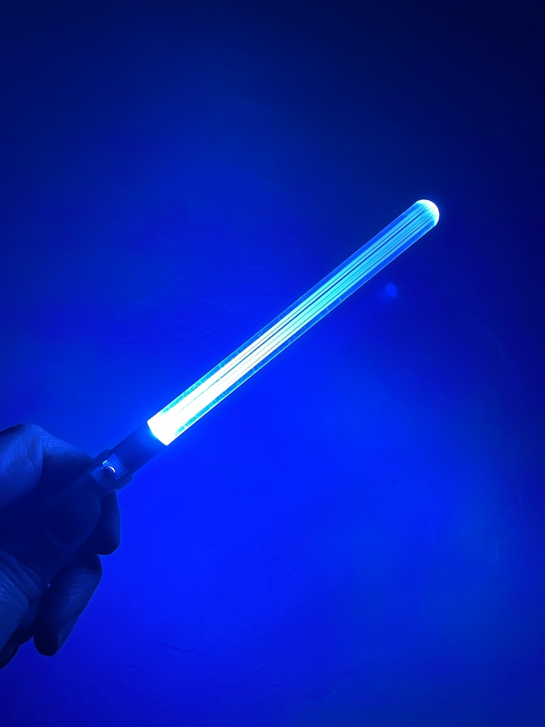 Peut inclure: Une baguette lumineuse bleue avec une lueur blanche. La baguette est tenue dans une main sur un fond bleu.