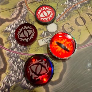 May include: A collection of game tokens featuring an eye design. The tokens are red, black, and silver, with the eye design in white. The tokens are on a game board with text that reads "PINNA GE" and "GON".