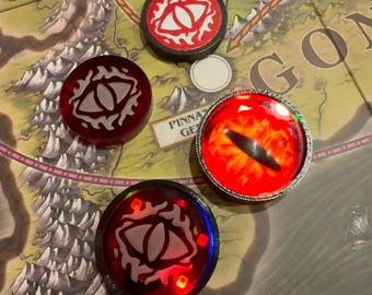 Peut inclure: Plusieurs jetons de jeu avec un motif d'œil. Les jetons sont rouges, noirs et argentés, avec le motif de l'œil en blanc. Les jetons sont sur un plateau de jeu avec le texte "PINNA GE" et "GON".