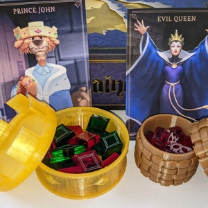 May include: Two golden treasure chests with lids open, revealing red and green gems inside. The chests are on a table with two cards in the background. The cards feature illustrations of Prince John and the Evil Queen.