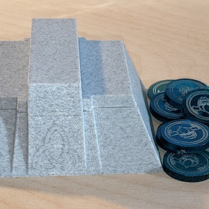 Pu&ograve; includere: Un pezzo da gioco di forma piramidale grigia con una pila di gettoni da gioco color verde acqua con un design stilizzato.