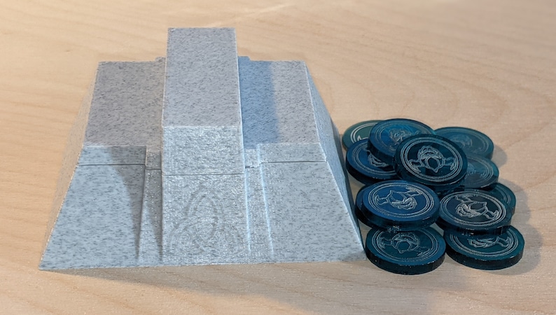 Pu&ograve; includere: Un pezzo da gioco di forma piramidale grigia con una pila di gettoni da gioco color verde acqua con un design stilizzato.