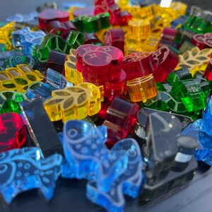 May include: A collection of colourful glass game pieces in various shapes, including leaves, fish, and abstract designs. The pieces are made of clear glass with different colours, including red, green, blue, and yellow.