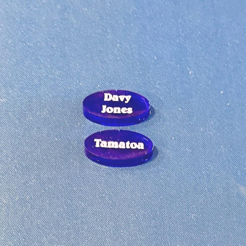 May include: Two oval, dark blue acrylic pieces with white text. The top piece reads "Davy Jones," and the bottom piece reads "Tamatoa." They are on a blue textured surface.