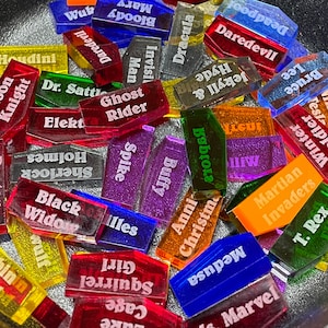 May include: A collection of colorful, rectangular acrylic tiles with various superhero and character names printed on them, including Daredevil, Dracula, Ghost Rider, Black Widow, and more.