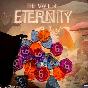 May include: The Vale of Eternity board game box with various game pieces. The game pieces are translucent and come in orange, purple, and blue. The game pieces have the numbers 1, 3, and 6 on them. The game box has the title "The Vale of Eternity".