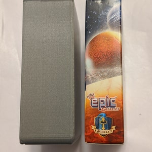 Pu&ograve; includere: Una scatola grigia accanto a una scatola con un'immagine colorata di pianeti e stelle. La scatola ha il testo "ultra tiny epic galaxies" e "Gamelyn Games".