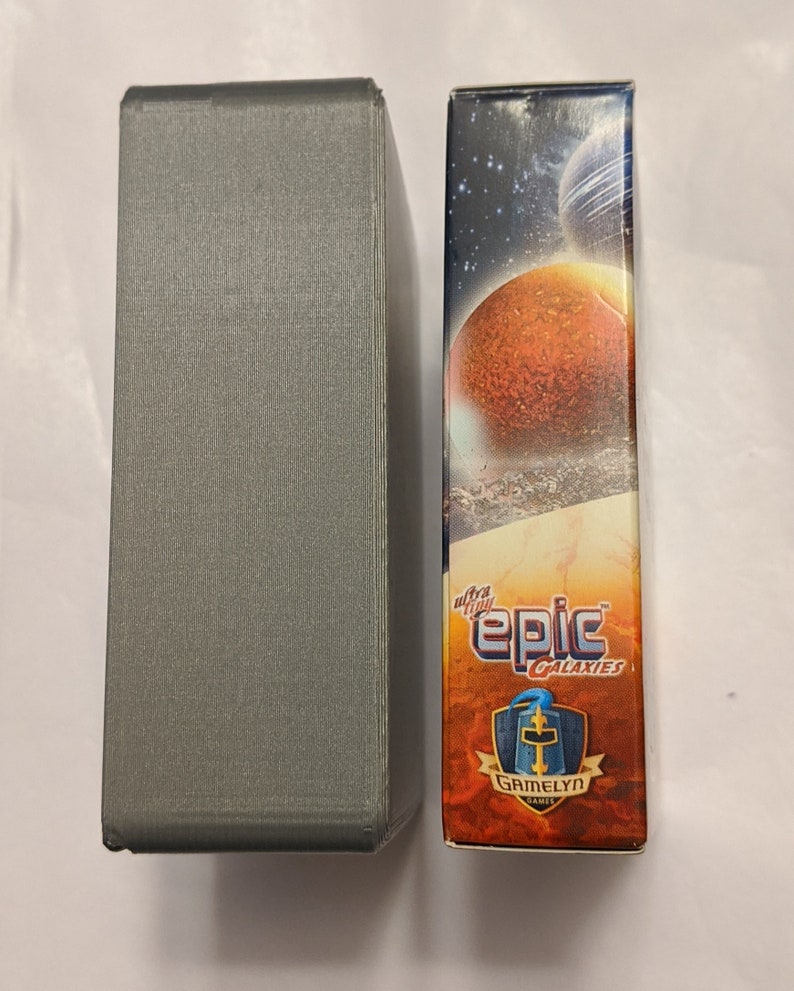 Pu&ograve; includere: Una scatola grigia accanto a una scatola con un'immagine colorata di pianeti e stelle. La scatola ha il testo "ultra tiny epic galaxies" e "Gamelyn Games".