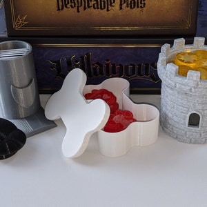 May include: A set of game pieces for a board game, including a gray tower with gold coins inside, a gray card holder, a white bone-shaped box with red tokens inside, and a black round token.