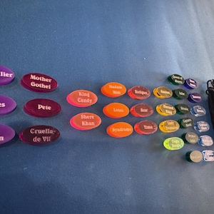 May include: A selection of oval game tokens, each bearing a character name such as "Hades" and "Cruella de Vil". The tokens are in shades of purple, red, orange, green, and blue. A black drawstring bag is also included in the image.