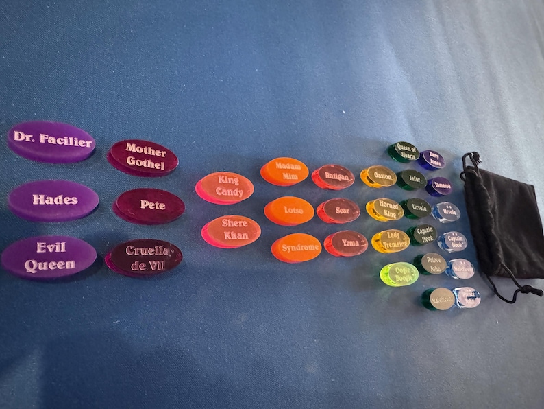 May include: A selection of oval game tokens, each bearing a character name such as "Hades" and "Cruella de Vil". The tokens are in shades of purple, red, orange, green, and blue. A black drawstring bag is also included in the image.