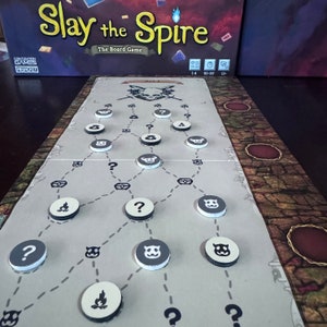 May include: The board game "Slay the Spire" is shown, with a game board displaying a path of circular tokens. The tokens feature question marks, skull and crossbones, and other symbols. The game box is visible in the background.