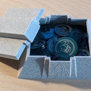 Pu&ograve; includere: Una scatola grigia testurizzata a forma di piramide con un coperchio incernierato. La scatola &egrave; aperta e contiene un mucchio di gettoni rotondi verdi con un design bianco. La scatola ha un design a nodo celtico sul fondo.