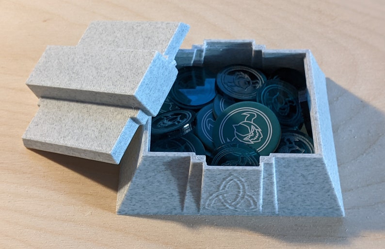 Pu&ograve; includere: Una scatola grigia testurizzata a forma di piramide con un coperchio incernierato. La scatola &egrave; aperta e contiene un mucchio di gettoni rotondi verdi con un design bianco. La scatola ha un design a nodo celtico sul fondo.