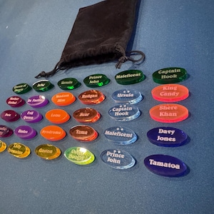 May include: A selection of brightly coloured, oval game pieces, each inscribed with character names such as "Ursula" and "Prince John". A black drawstring bag is positioned above the pieces on a blue surface.
