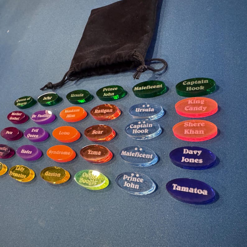 May include: A selection of brightly coloured, oval game pieces, each inscribed with character names such as "Ursula" and "Prince John". A black drawstring bag is positioned above the pieces on a blue surface.