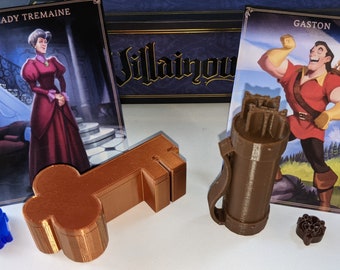 May include: A brown key-shaped box and a brown cylindrical box with a strap. The boxes are on a white surface. There are two cards in the background, one with the text "Lady Tremaine" and the other with the text "Gaston".