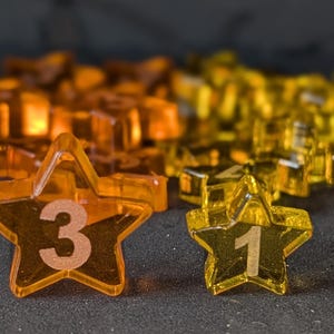 May include: Close-up of star-shaped game pieces in shades of orange and yellow. Two pieces are in focus, one with the number 3 and the other with the number 1. The background shows a blurred pile of similar pieces.