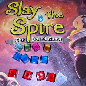 May include: A close-up of a game board with the title "Slay the Spire: The Board Game".  The board is surrounded by colorful glass tokens in red, blue, green, and purple.