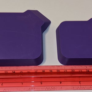 Two purple stackable token trays shown upside down alongside a red ruler so you can measure the square tray area and built-in bagging funnel details.