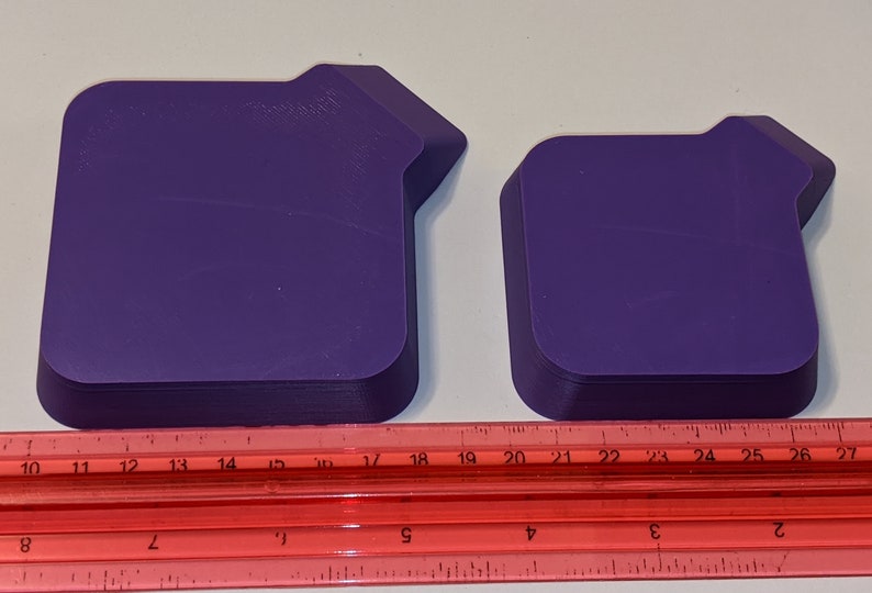 Two purple stackable token trays shown upside down alongside a red ruler so you can measure the square tray area and built-in bagging funnel details.