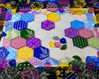 May include: A close-up of a game board with hexagonal tiles featuring various patterns and colors, including green, blue, purple, pink, and yellow. The tiles are arranged in a honeycomb pattern on a white background. There are also several smaller, colorful tokens scattered around the board.