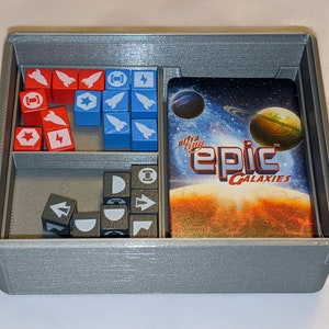 Pu&ograve; includere: Un organizzatore grigio stampato in 3D con scomparti per i pezzi di gioco e una carta. La carta ha un tema spaziale e dice "ultra tiny epic galaxies".