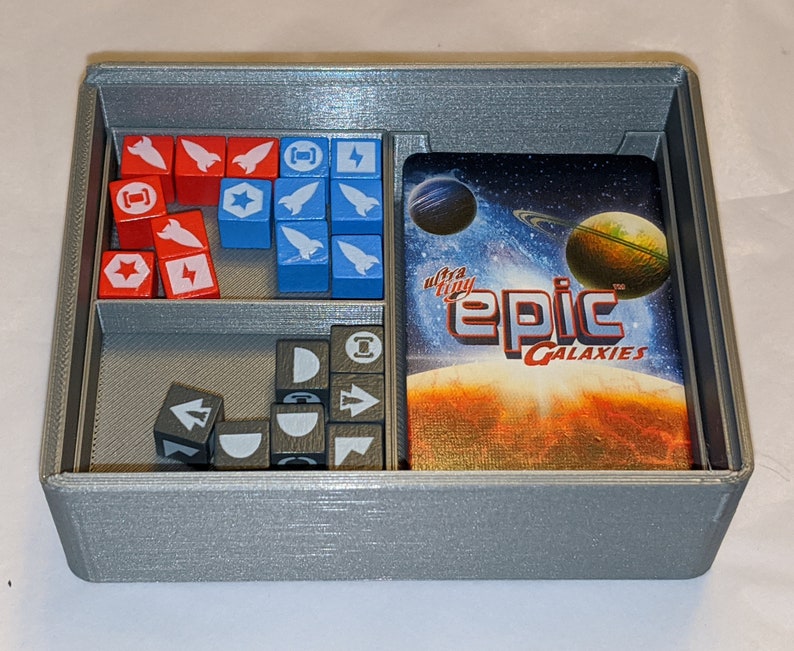 Pu&ograve; includere: Un organizzatore grigio stampato in 3D con scomparti per i pezzi di gioco e una carta. La carta ha un tema spaziale e dice "ultra tiny epic galaxies".
