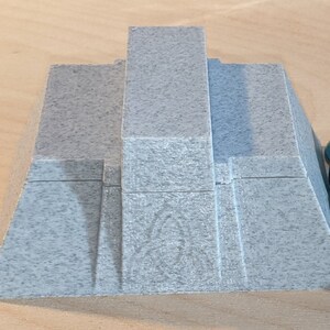 Pu&ograve; includere: Una scatola grigia a forma di piramide con una superficie testurizzata. La scatola &egrave; realizzata in plastica e ha un coperchio che si apre per rivelare un vano all'interno. La scatola &egrave; appoggiata su una superficie di legno.