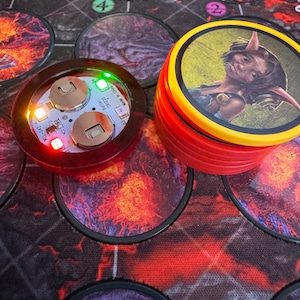 May include: A board game setup featuring a red and yellow stack of character tokens with a fantasy illustration. A lit, round, red electronic component with green and white lights sits nearby. The game board has a dark, fiery design.