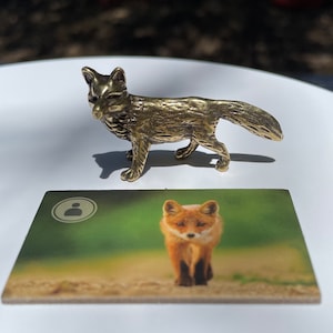 May include: A gold-colored metal figurine of a fox standing on a white surface. The fox is facing the left side of the image. A card with a picture of a red fox is in the background.