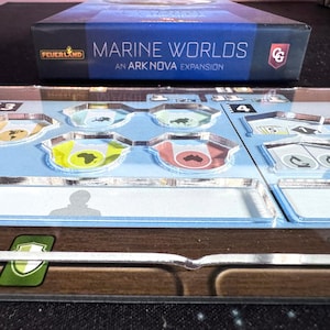 May include: A board game box titled "MARINE WORLDS: AN ARK NOVA EXPANSION" sits atop a game board. The box is dark blue with white text and a logo. The game board has light blue sections with clear plastic overlays and various colorful game pieces.