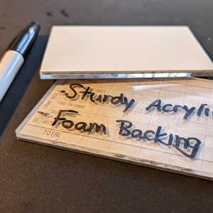 May include: A clear acrylic sheet with a grid pattern and the words "Sturdy Acrylic Foam Backing" written in black marker.