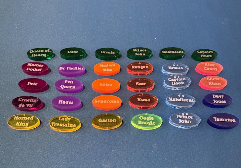 May include: A selection of colourful, oval acrylic tokens, each inscribed with a different name. The tokens are in various colours, including green, purple, orange, red, and blue. The names are in white, and include characters like "Ursula" and "Maleficent".