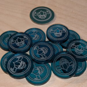 Pu&ograve; includere: Un mucchio di gettoni da gioco blu-verde con un design argentato di una creatura con corna e zanne.