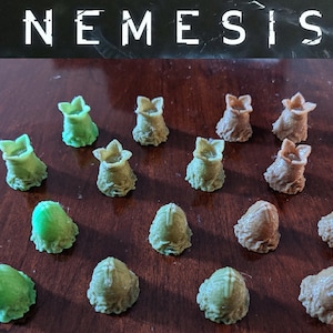 May include: A collection of miniature game pieces, including green and tan alien eggs and pod-like figures. The word "NEMESIS" is printed in white at the top of the image. The pieces are arranged on a dark wooden surface.
