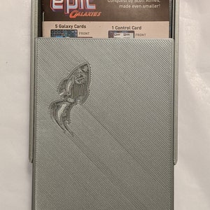 Pu&ograve; includere: Una scatola di plastica grigia con un design a forma di pesce argentato sul davanti. La scatola contiene un set di carte per il gioco "Epic Galaxies: A Game of Intergalactic Conquest by Scott Almes, made even smaller!" Le carte includono 5 carte Galaxy e 1 carta di controllo.