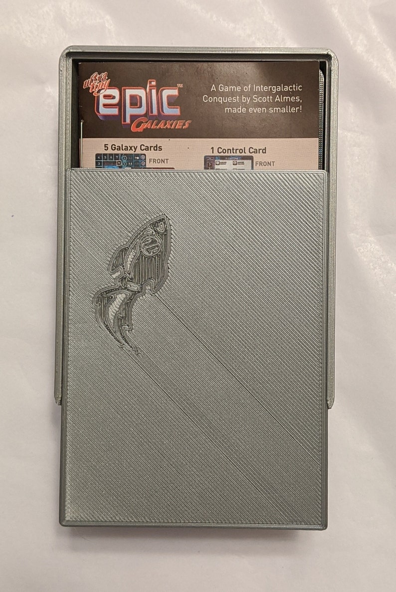 Pu&ograve; includere: Una scatola di plastica grigia con un design a forma di pesce argentato sul davanti. La scatola contiene un set di carte per il gioco "Epic Galaxies: A Game of Intergalactic Conquest by Scott Almes, made even smaller!" Le carte includono 5 carte Galaxy e 1 carta di controllo.