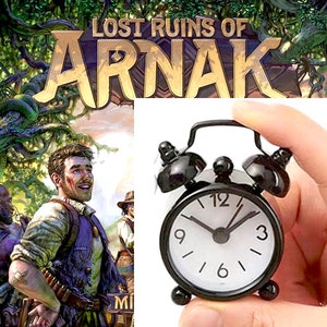 May include: A black alarm clock with white face and black hands. The clock is set to 12:00. The clock is being held in a hand.